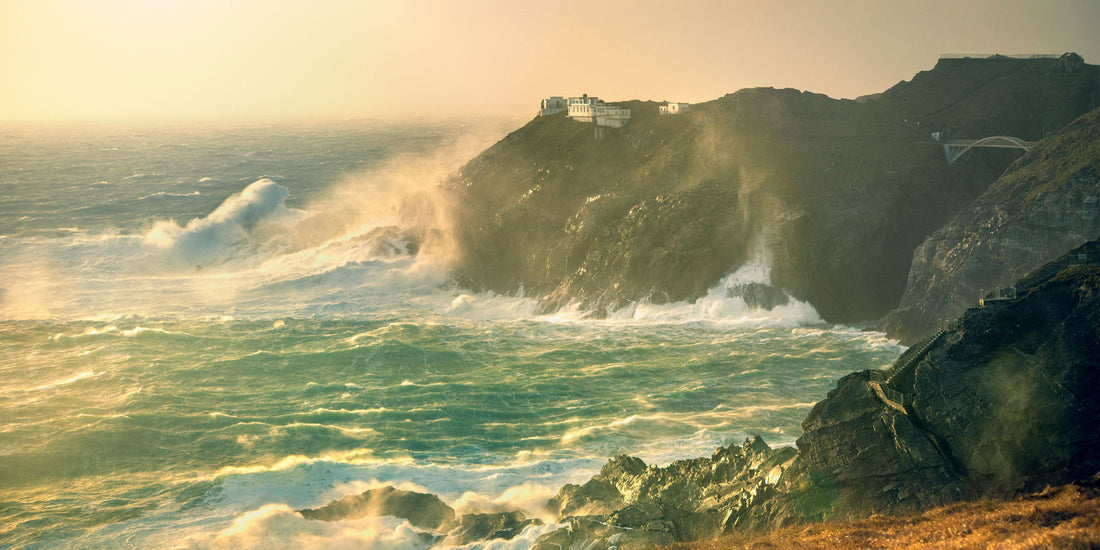 Raging ocean waves crashing against a cliff with a lighthouse in the background.
